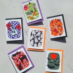 May include: Six Halloween-themed greeting cards with various illustrations. The cards feature mushrooms, ghosts, flowers, cats with eyes, and bats with a red sun. Each card has a unique design and colour scheme, with black, orange, and purple borders.