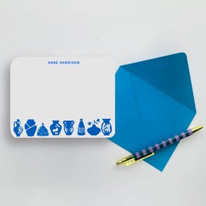 Peut inclure: Une carte de correspondance rectangulaire blanche aux coins arrondis avec le texte bleu ANNE HARRISON et une rangée d'illustrations de vases bleus en bas, accompagnée d'une enveloppe bleu vif et d'un stylo à bille rayé violet et vert.