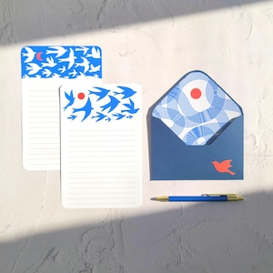 Puede incluir: Juego de papelería con diseño de pájaros azules y blancos. Incluye dos blocs de notas con páginas rayadas y un sobre a juego. Un bolígrafo azul con detalles dorados descansa cerca. El sobre tiene una pegatina de pájaro rojo.