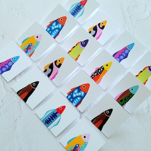 Puede incluir: Una colección de tarjetas cuadradas blancas, cada una con una ilustración colorida y estilizada de un pez. Los diseños de los peces varían en color y patrón, con tonos vibrantes de azul, naranja, amarillo, rosa y verde. Los peces están representados con un estilo artístico minimalista.