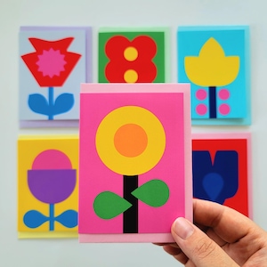 May include: A selection of vibrant floral greeting cards. Each card showcases a distinct, stylized flower design in bold colours including red, yellow, blue, and pink. The cards are displayed against a white backdrop, with one card held by a hand.