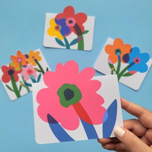 Peut inclure: Quatre cartes de vœux rectangulaires présentant des illustrations florales colorées au style plat. La carte au premier plan affiche une grande fleur rose avec un centre vert et des feuilles bleues. Les autres cartes montrent des fleurs rouges, jaunes, orange et bleues.
