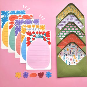 May include: A collection of stationery with floral designs. The set includes lined notepads with flower illustrations and patterned envelopes in green hues. Small flower-shaped cutouts are scattered below the notepads.
