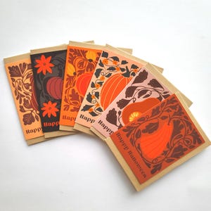 以下が含まれることがあります： 茶色のクラフト紙の裏地が付いた、ハロウィーンのグリーティングカード6枚のコレクション。各カードには、オレンジ、茶色、ピンクの色合いの、カボチャと花の要素の異なるデザインが施されています。「Happy Halloween」の文字がカードに印刷されています。