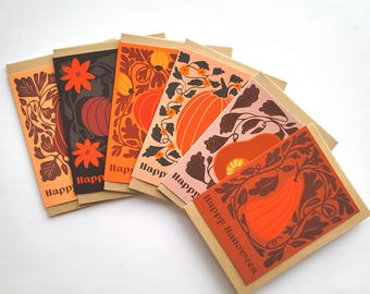 Peut inclure: Une collection de six cartes de vœux d'Halloween avec un dos en papier kraft brun. Chaque carte présente un design différent de citrouilles et d'éléments floraux dans des tons orange, marron et rose. Les mots "Happy Halloween" sont imprimés sur les cartes.