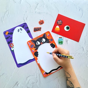 May include: A Halloween stationery set with a black cat, ghost, and pumpkin design. Includes a red envelope with stickers, a jar with a frog, and a pen. Ideal for seasonal notes and greetings. The pen is being held by a person.