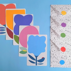 Puede incluir: Un conjunto de tarjetas de arte floral coloridas con diseños abstractos en naranja, azul, rosa y rojo. También se incluye un juego de sobres blancos moteados con cierres de puntos de colores. Sobre un fondo azul claro.