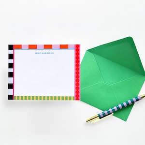 Op de afbeelding: Een witte rechthoekige briefkaart met de naam ANNE HARRISON in blauw gedrukt, voorzien van een rand met zwart-witte strepen, oranje en roze blokken, rode stippen en groene strepen, vergezeld van een groene envelop en een gestreepte pen.