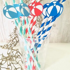 May include: A glass filled with red, blue, and turquoise striped paper straws with blue and red beach ball cutouts attached to the ends.