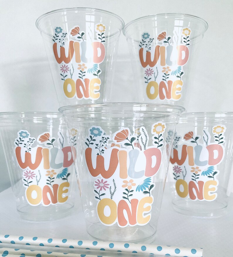May include: Set of six clear plastic cups with colorful "Wild One" stickers featuring floral designs. The stickers are in shades of yellow, orange, blue, and pink.