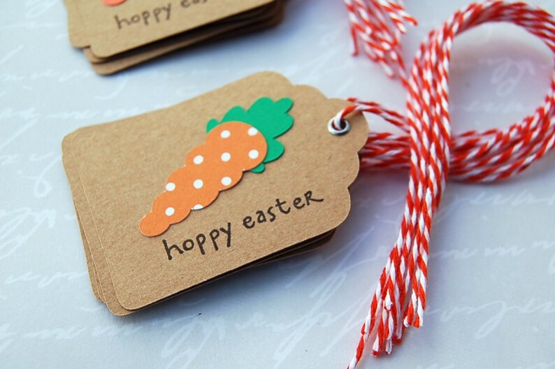 May include: A stack of brown gift tags with a white string attached. The tags are decorated with a paper carrot with white polka dots and green leaves. The text "happy easter" is written on the tag.