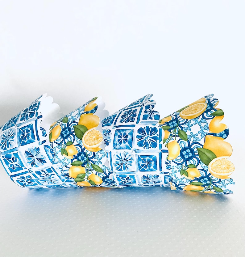 May include: Decorative paper crown featuring a blue and white tile pattern with yellow lemons and green leaves. The crown has a scalloped edge and is designed to be worn. The design is reminiscent of Mediterranean decor.