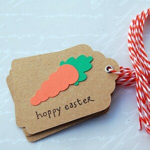 May include: A brown paper gift tag with a green and orange carrot cutout and the text "hoppy easter". The tag is tied with a red and white baker's twine.