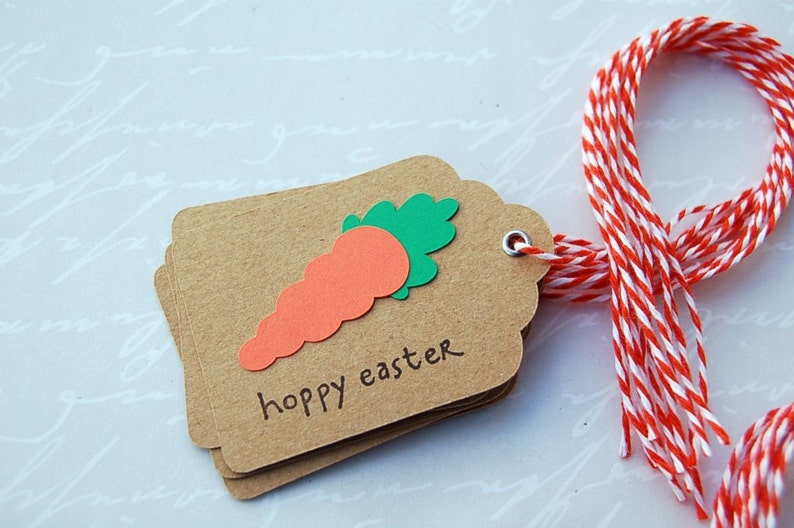 May include: A brown paper gift tag with a green and orange carrot cutout and the text "hoppy easter". The tag is tied with a red and white baker's twine.
