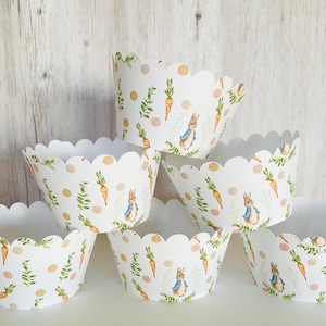 May include: White cupcake liners with a scalloped edge feature a repeating pattern of a bunny rabbit, carrots, and green leaves. The bunny is wearing a blue jacket and has brown fur.