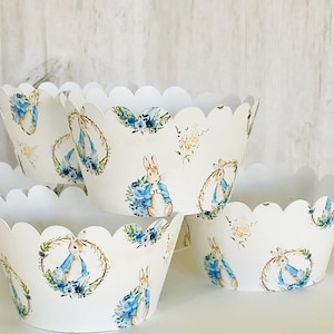 May include: White cupcake liners with a scalloped edge and a repeating pattern of a blue bunny in a blue jacket, blue flowers, and a gold wreath.
