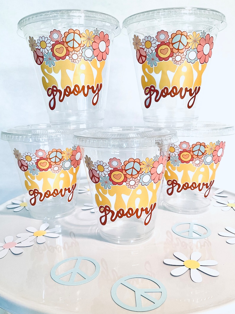Peut inclure: Cinq gobelets en plastique transparent avec un motif floral et un signe de paix et le texte "Stay Groovy" en lettres jaunes et brunes. Les gobelets sont sur une surface blanche avec des marguerites en papier et des signes de paix.