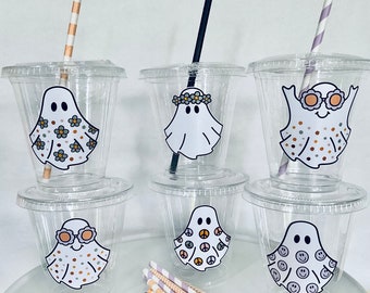 May include: Six clear plastic cups with lids and straws decorated with ghost designs. The ghosts have different patterns, including flowers, peace signs, and smiley faces. The straws are black and striped.