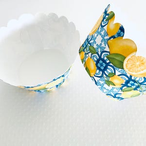 May include: Two white cupcake liners with scalloped edges. One liner is empty, the other has a decorative pattern of lemons, blue floral designs, and green leaves. The liners are designed for baking.