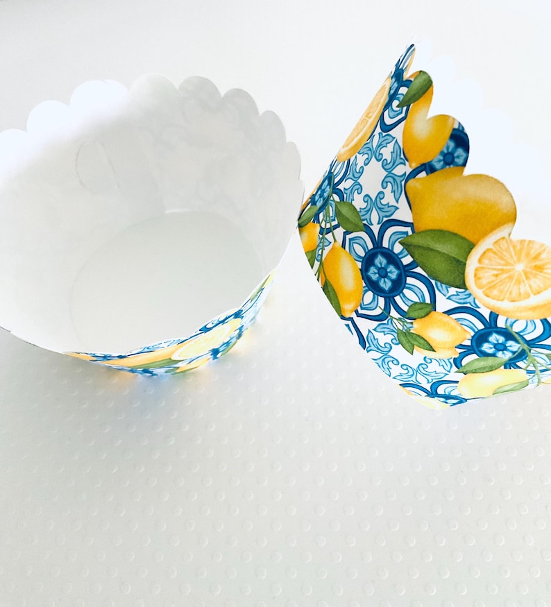 May include: Two white cupcake liners with scalloped edges. One liner is empty, the other has a decorative pattern of lemons, blue floral designs, and green leaves. The liners are designed for baking.