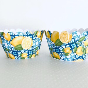 May include: Two paper cupcake liners with a scalloped edge. The liners feature a repeating pattern of yellow lemons, green leaves, and blue floral designs. The background is white.