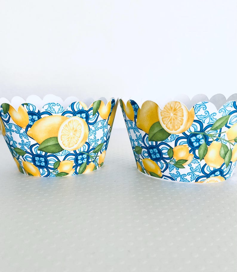 May include: Two paper cupcake liners with a scalloped edge. The liners feature a repeating pattern of yellow lemons, green leaves, and blue floral designs. The background is white.