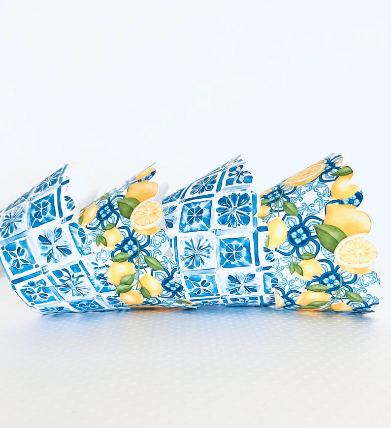 May include: Decorative paper crown with a blue and white tile pattern and lemon illustrations. The crown is made of folded paper and has a scalloped edge. The lemons are yellow with green leaves.