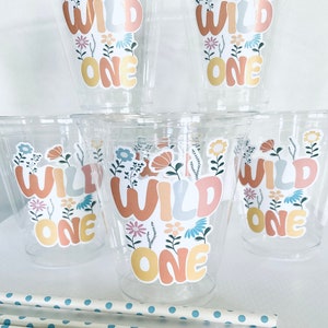 May include: Set of six clear plastic cups with floral and wild one stickers. The cups are stacked on top of each other. There are also blue and white striped straws in the image.