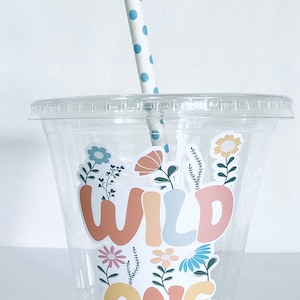 May include: A clear plastic cup with a straw and a sticker that says "WILD ONE" in colorful letters with flowers and leaves around the text.