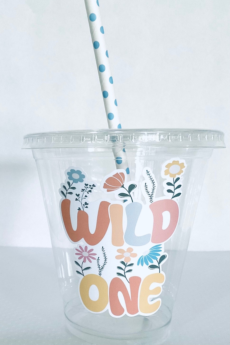 May include: A clear plastic cup with a straw and a sticker that says "WILD ONE" in colorful letters with flowers and leaves around the text.