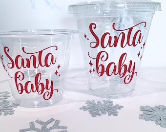 May include: Clear plastic cups with red text that reads "Santa baby". One cup has a lid. Silver glitter snowflake decorations are scattered around the cups. The background has a light blue snowflake design.