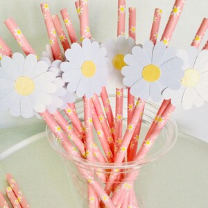 May include: Pink and white striped paper straws with white daisy flower cutouts. The straws are in a clear plastic cup.