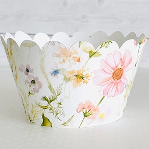 May include: A white paper cupcake liner with a scalloped edge, adorned with a watercolor floral pattern. The design features pink, yellow, and blue flowers with green foliage, creating a delicate and charming aesthetic.