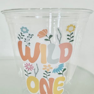 May include: A clear plastic cup with a sticker that says "Wild One" in colorful letters surrounded by flowers.