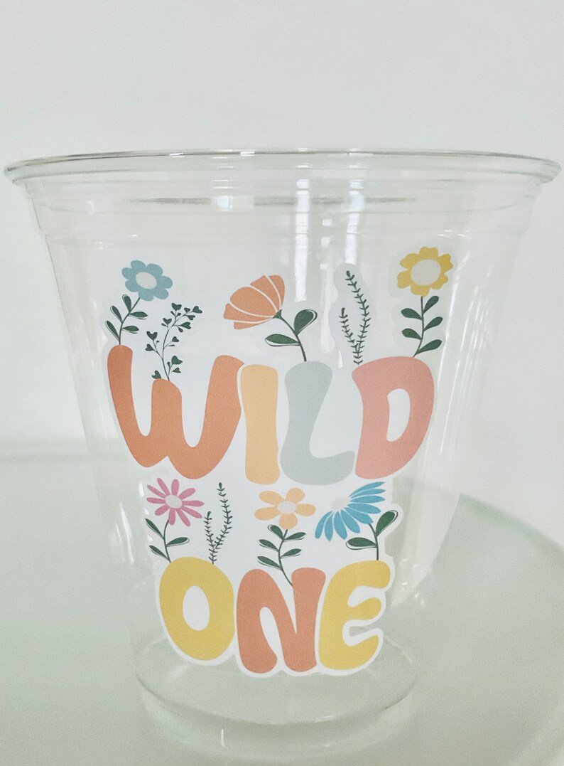May include: A clear plastic cup with a sticker that says "Wild One" in colorful letters surrounded by flowers.