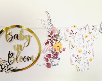 May include: A gold circle banner with the text "Baby in Bloom" and floral accents. The banner is strung with a white string and includes a floral design and a baby onesie design.