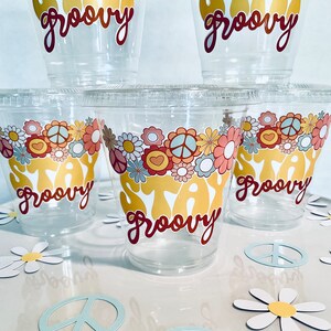 Peut inclure: Cinq gobelets en plastique transparent avec un motif "Stay Groovy" en or et rouge. Les gobelets sont d&eacute;cor&eacute;s de signes de paix et de fleurs. Les gobelets sont pos&eacute;s sur une surface blanche avec des signes de paix et des marguerites en papier.