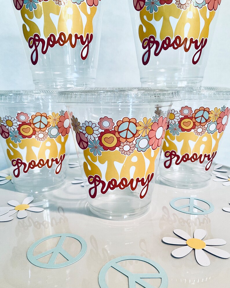 Peut inclure: Cinq gobelets en plastique transparent avec un motif "Stay Groovy" en or et rouge. Les gobelets sont d&eacute;cor&eacute;s de signes de paix et de fleurs. Les gobelets sont pos&eacute;s sur une surface blanche avec des signes de paix et des marguerites en papier.