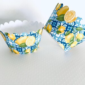 May include: Two cupcake liners with a scalloped edge. The liners feature a pattern of lemons, green leaves, and blue and white decorative tiles. One liner is upright, and the other is tilted.