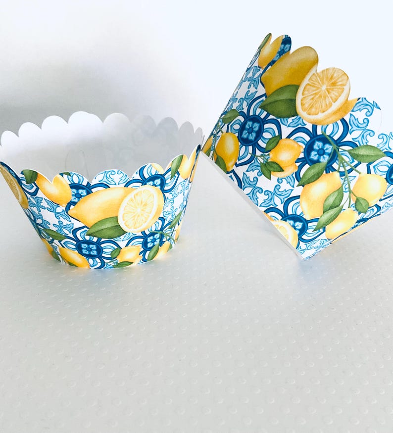 May include: Two cupcake liners with a scalloped edge. The liners feature a pattern of lemons, green leaves, and blue and white decorative tiles. One liner is upright, and the other is tilted.