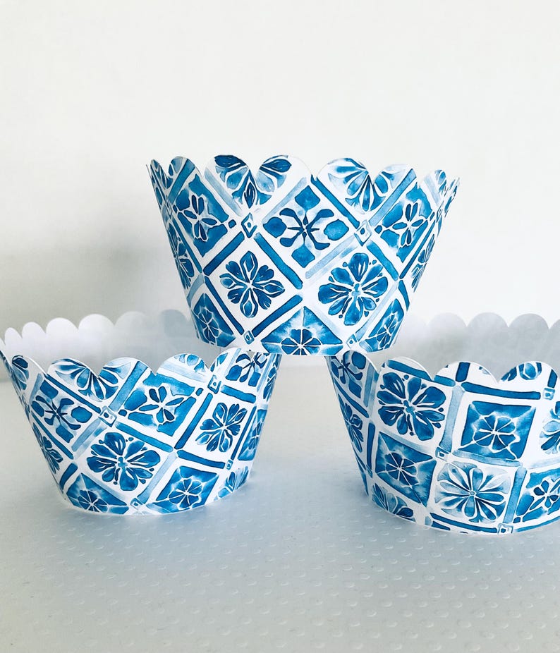 May include: Three blue and white patterned cupcake liners with a scalloped edge. The design features a floral and geometric pattern in shades of blue on a white background. The liners are stacked on top of each other.