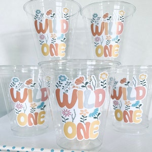 May include: Set of six clear plastic cups with colorful "Wild One" stickers featuring floral designs. The stickers are in shades of yellow, orange, blue, and pink.
