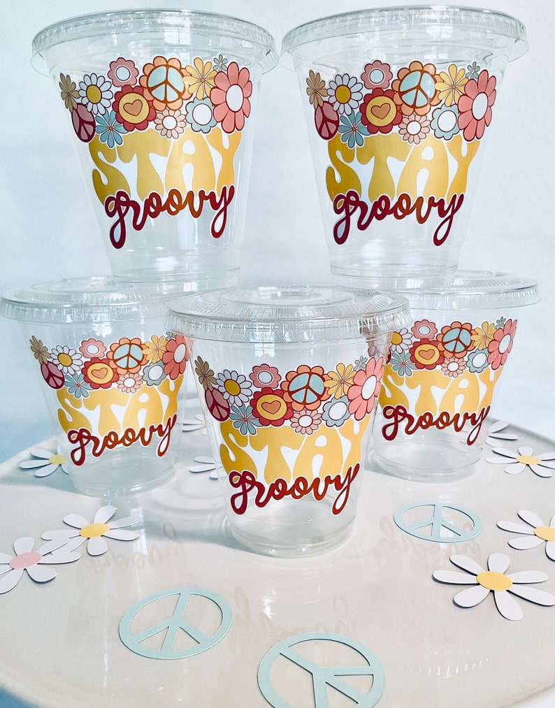 Peut inclure: Cinq gobelets en plastique transparent avec un motif floral et le texte "Stay Groovy" en jaune et rouge. Les gobelets sont sur une surface blanche avec des marguerites en papier blanc.