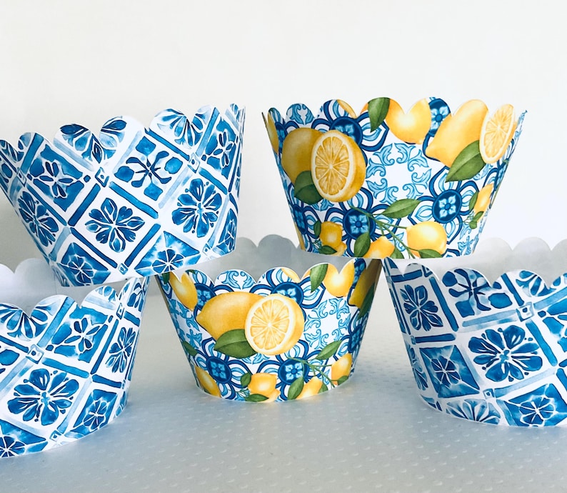May include: A collection of decorative cupcake liners. Some liners feature a blue and white tile pattern, while others showcase lemons and green leaves against a blue and white background. The liners have a scalloped edge.