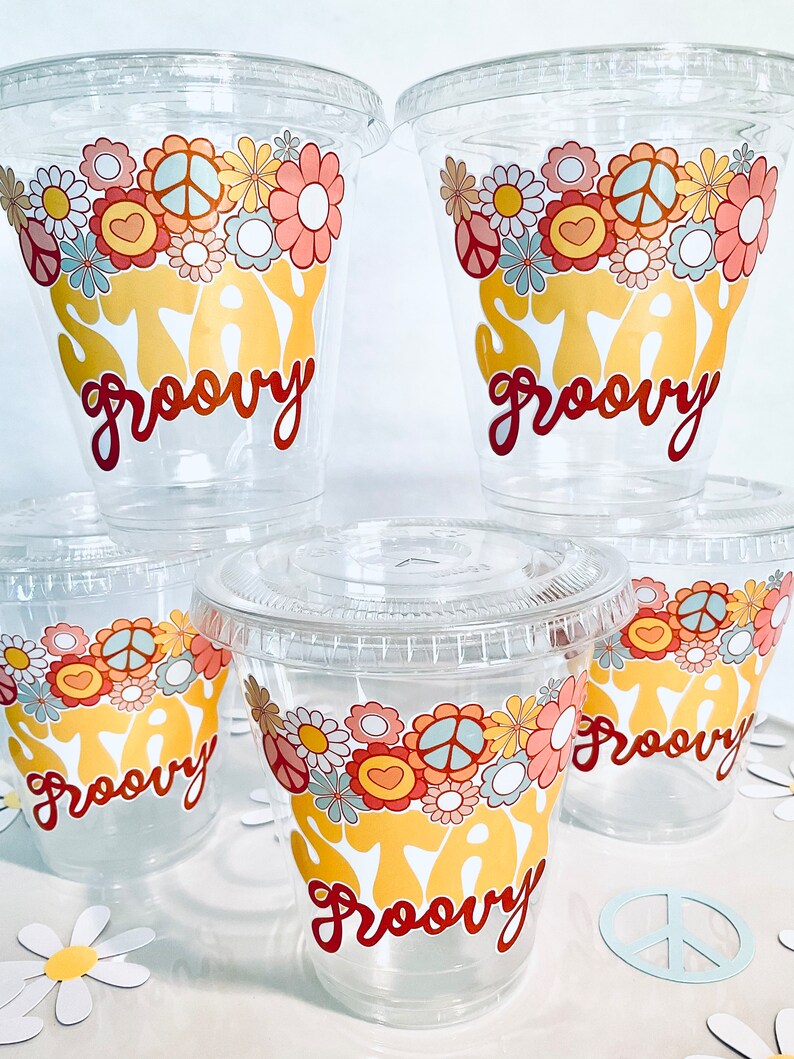 Peut inclure: Quatre gobelets en plastique transparent avec couvercles, chacun d&eacute;cor&eacute; d'un motif floral color&eacute; et du texte "Stay Groovy" en lettres jaunes et rouges.
