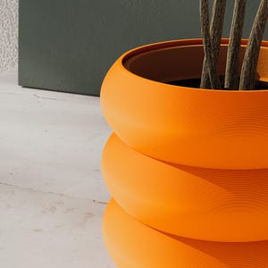 May include: A bright orange, cylindrical plant pot with a ribbed, stacked-ring texture. The exterior features a matte finish, and the interior is dark, containing the base of a plant with thin, brown, woody stems extending upward from the center.