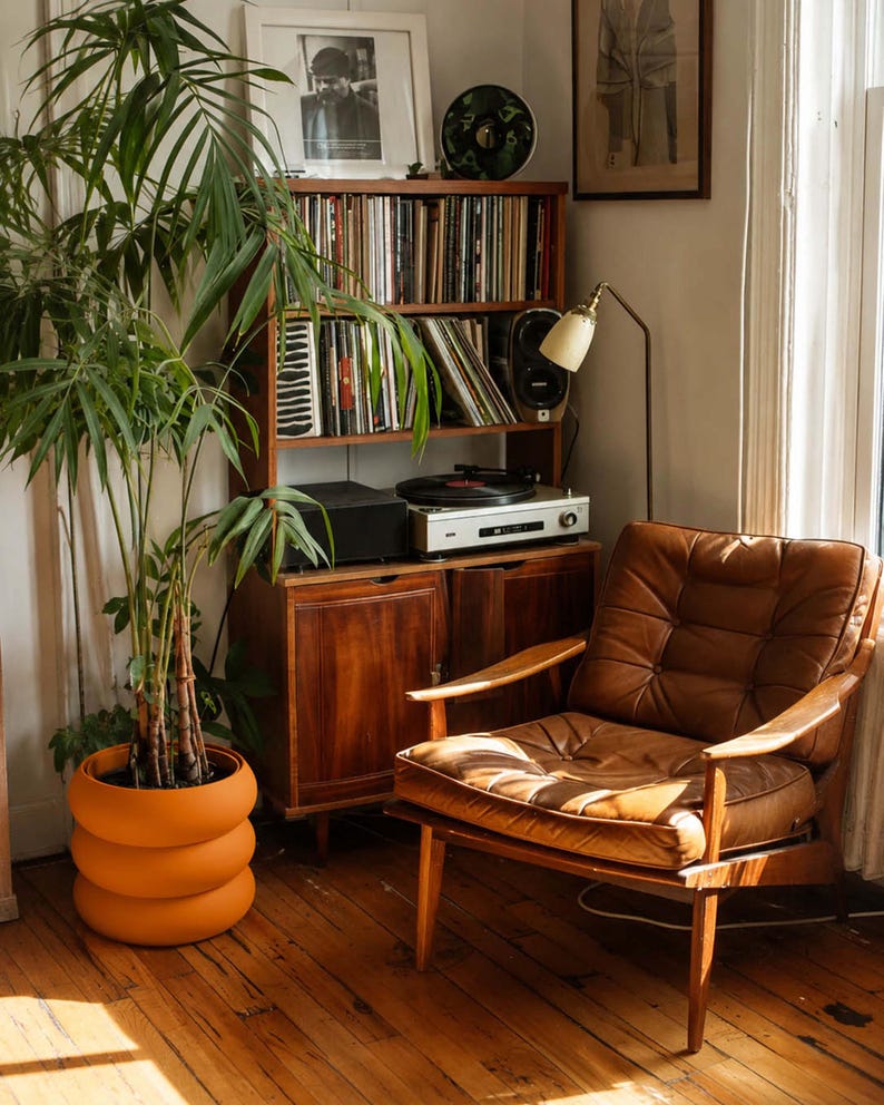 May include: A tall green palm plant in a bright orange, three-tiered, rounded planter sits next to a brown leather armchair with wooden armrests. Behind the chair is a wooden shelving unit holding vinyl records, a turntable, and a lamp.