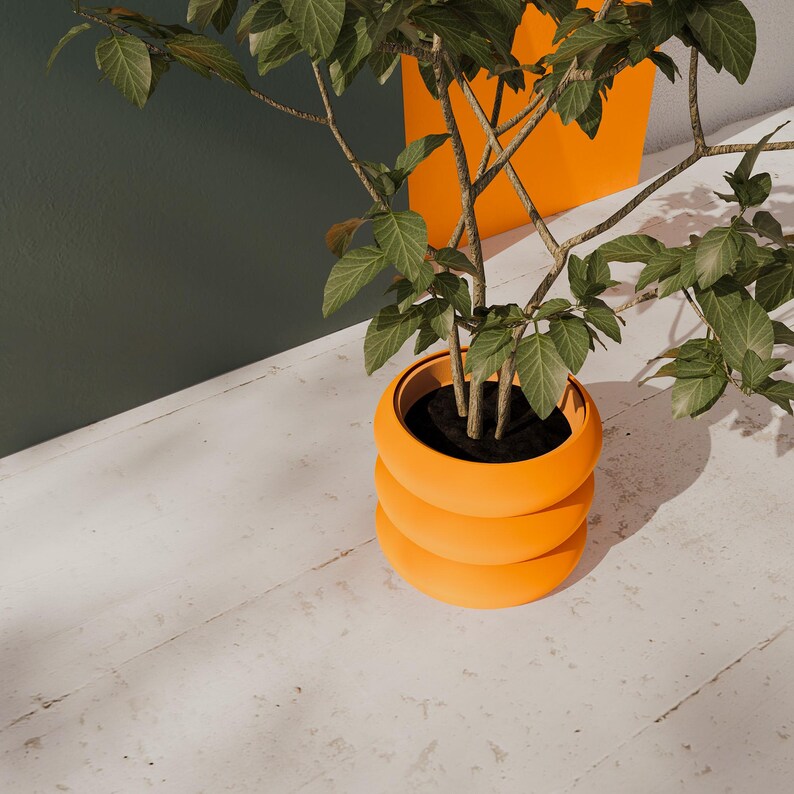 May include: A bright orange, three-tiered, rounded planter pot containing a green leafy plant with thin, woody stems. The planter has a smooth, matte finish and a wide, circular opening filled with dark soil, showcasing a vibrant, modern aesthetic.