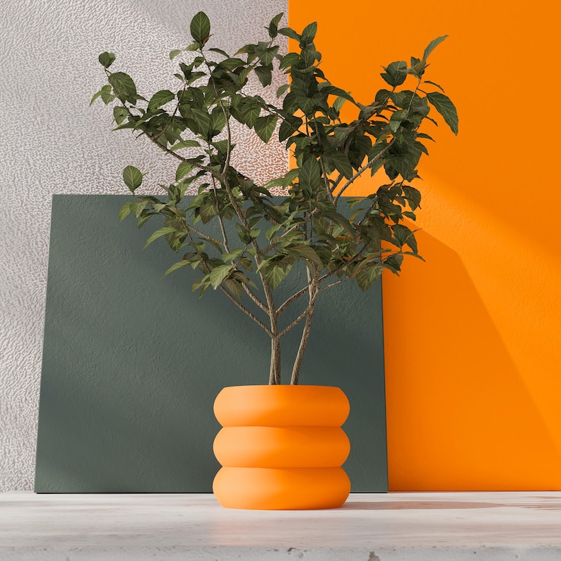 May include: A vibrant orange, cylindrical plant pot with three distinct, rounded horizontal ridges stacked vertically. The pot contains a leafy green plant with thin, branching stems and numerous small, pointed leaves extending upwards.