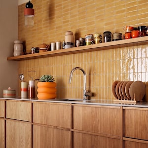 May include: A kitchen featuring light wood cabinets, a white countertop, and a backsplash of glossy mustard yellow rectangular tiles. A wooden shelf holds various ceramic mugs and containers, while a silver faucet sits above the sink.